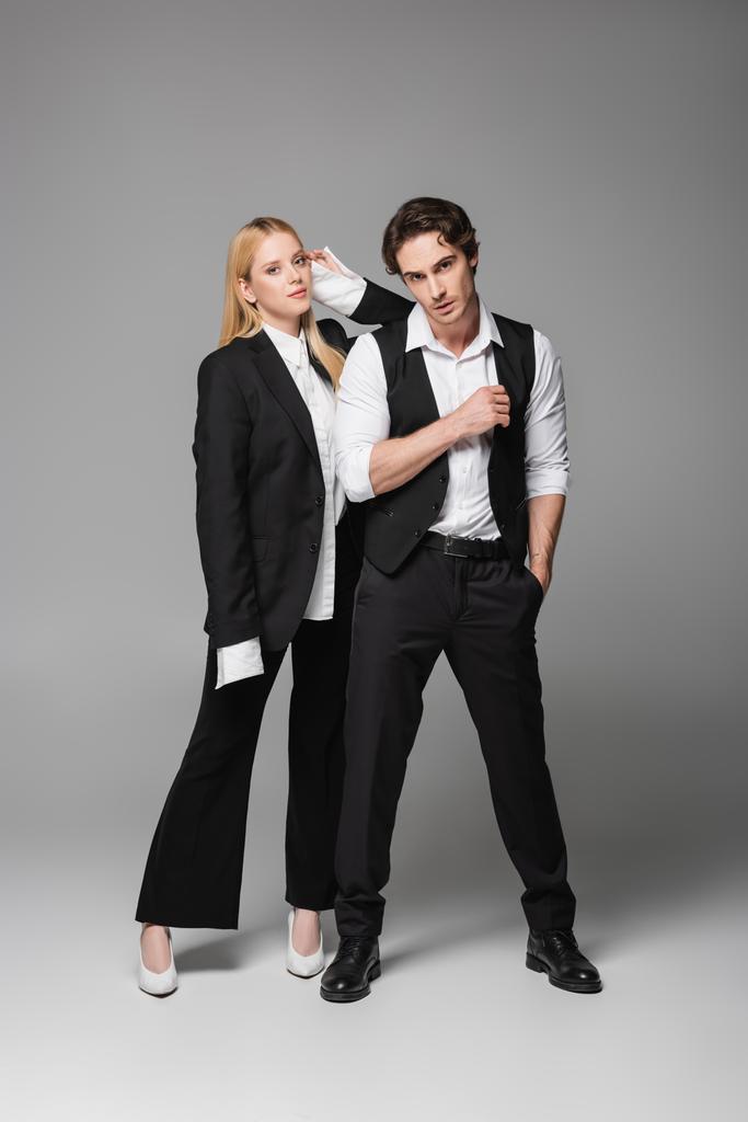 Ansicht des stilvollen Paares in eleganter Kleidung mit Blick auf die Kamera auf grau - Foto, Bild