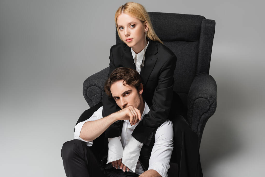 blonde woman in armchair looking at camera while embracing stylish man sitting on grey - Photo, Image