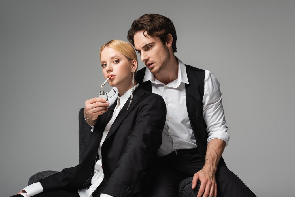 stijlvolle man in elegante kleren lichter sigaret van blonde vrouw geïsoleerd op grijs - Foto, afbeelding