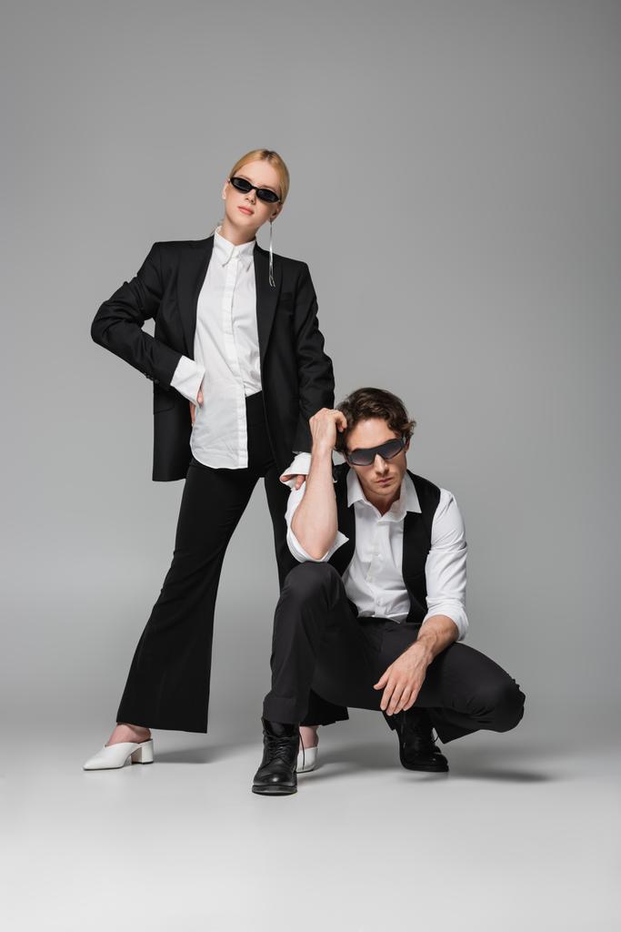 blonde woman in black suit standing with hand on hip near elegant man posing on grey - Photo, Image