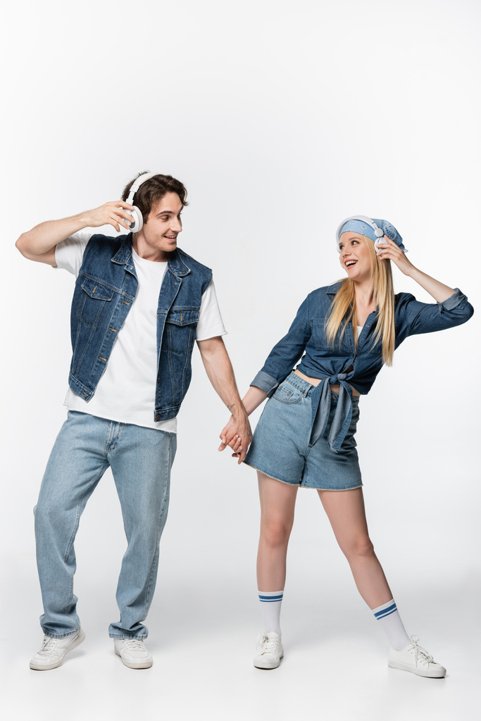 cheerful couple in denim clothing and headphones holding hands while listening music on white - Photo, Image