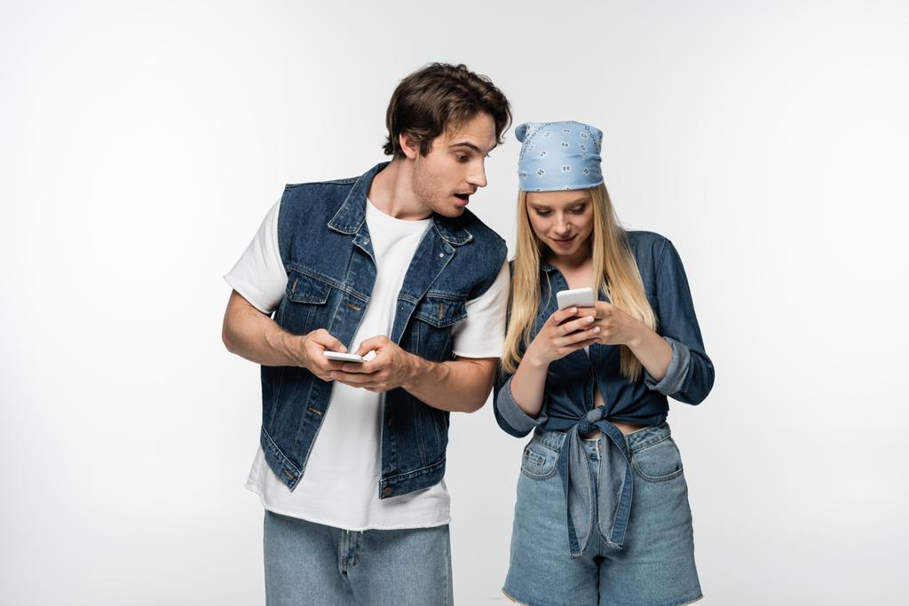 изумленный мужчина в джинсовой одежде смотрит на женщину с помощью мобильного телефона, изолированного на белом - Фото, изображение