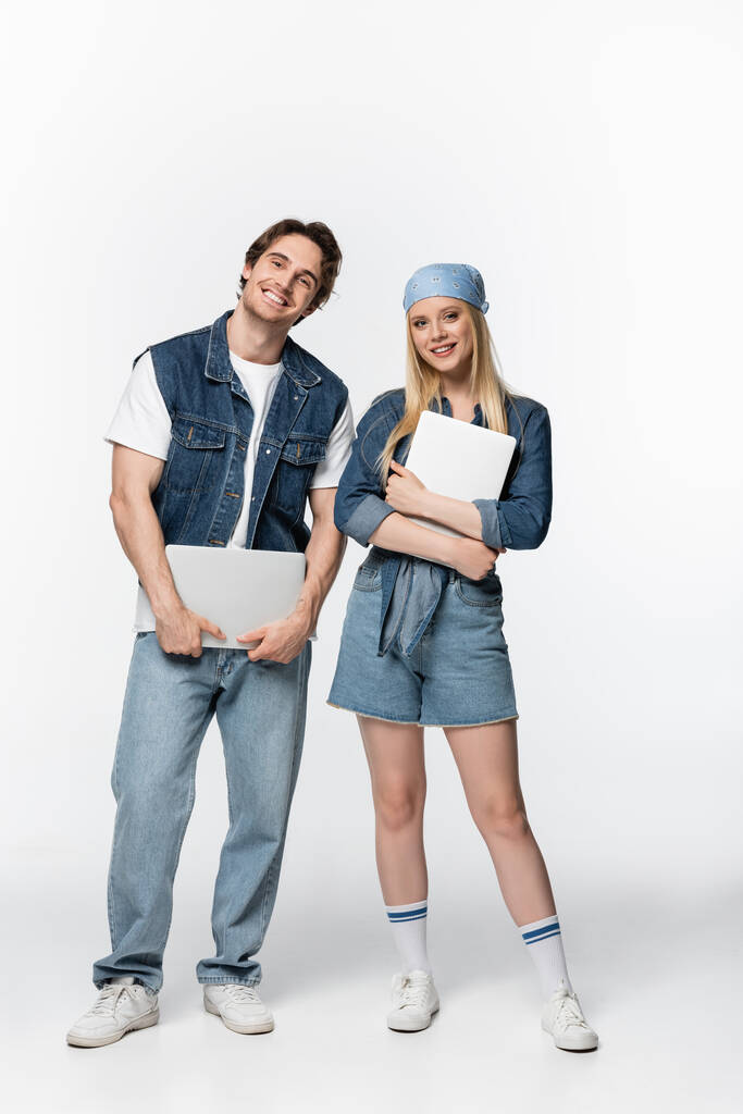 full length view of pleased and stylish couple with laptops smiling at camera on white - Photo, Image