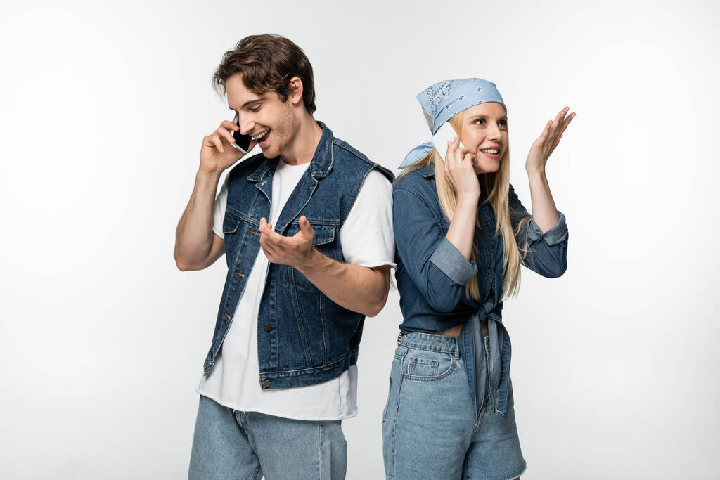 радостная пара в модной джинсовой одежде разговаривает на мобильных телефонах изолированных на белом - Фото, изображение