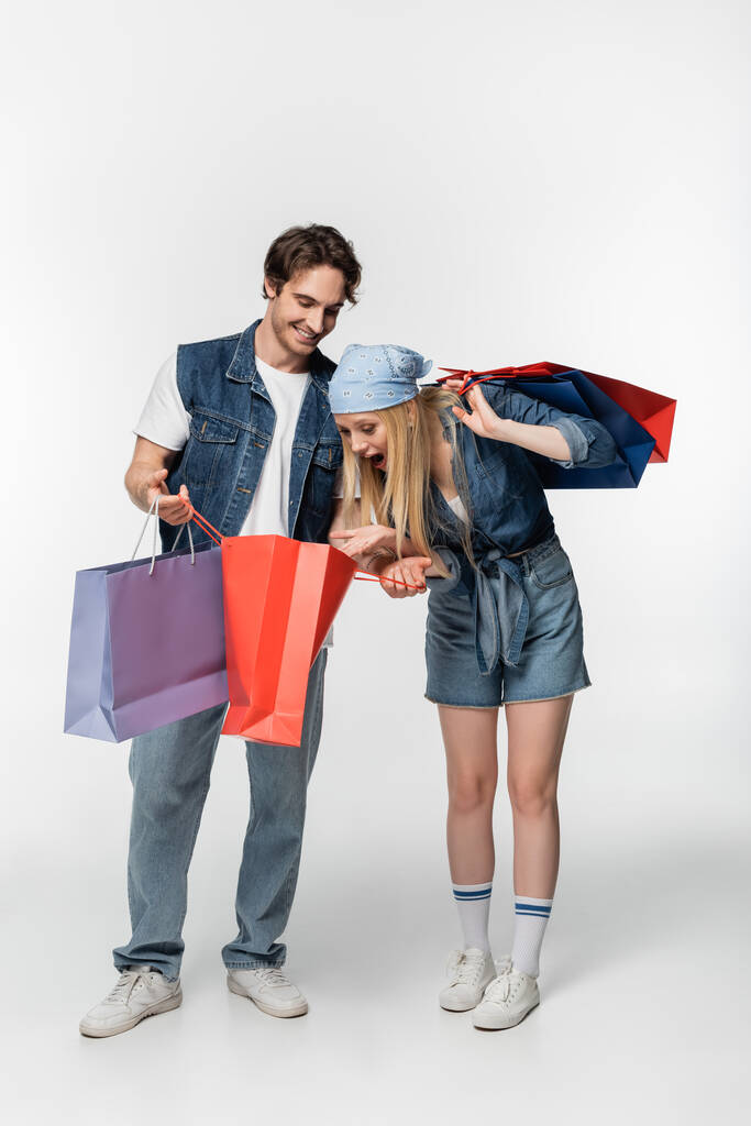 mulher espantada olhando no saco de compras em mãos de namorado elegante no branco - Foto, Imagem