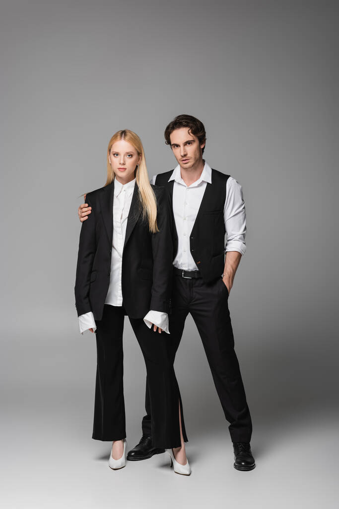 stijlvolle man staan met de hand in zak in de buurt van blonde vrouw op grijs - Foto, afbeelding
