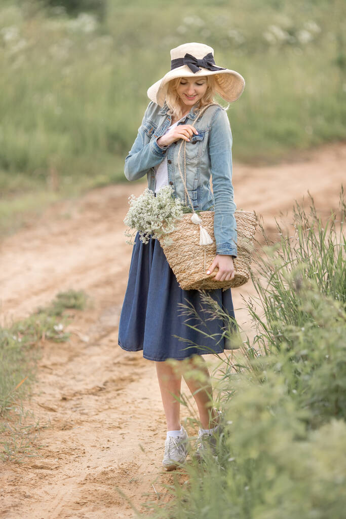 egy fiatal szőke nő kék szemekkel szalmakalapban és farmer ruhában egy szalmazsákot tart virágokkal a parkban egy nyári napon. A romantika fogalma - Fotó, kép