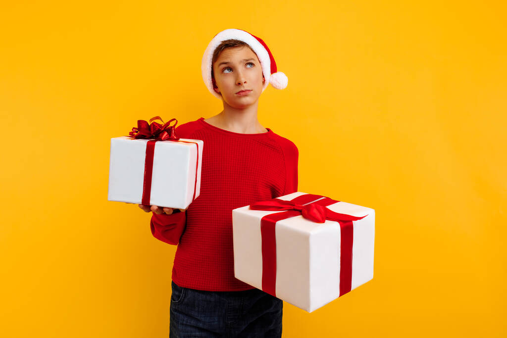 Garçon pensif dans un chapeau de Noël et un cadeau dans ses mains. La pensée pense à ce qu'il y a dans les cadeaux. Sur fond jaune. Concept de Noël - Photo, image