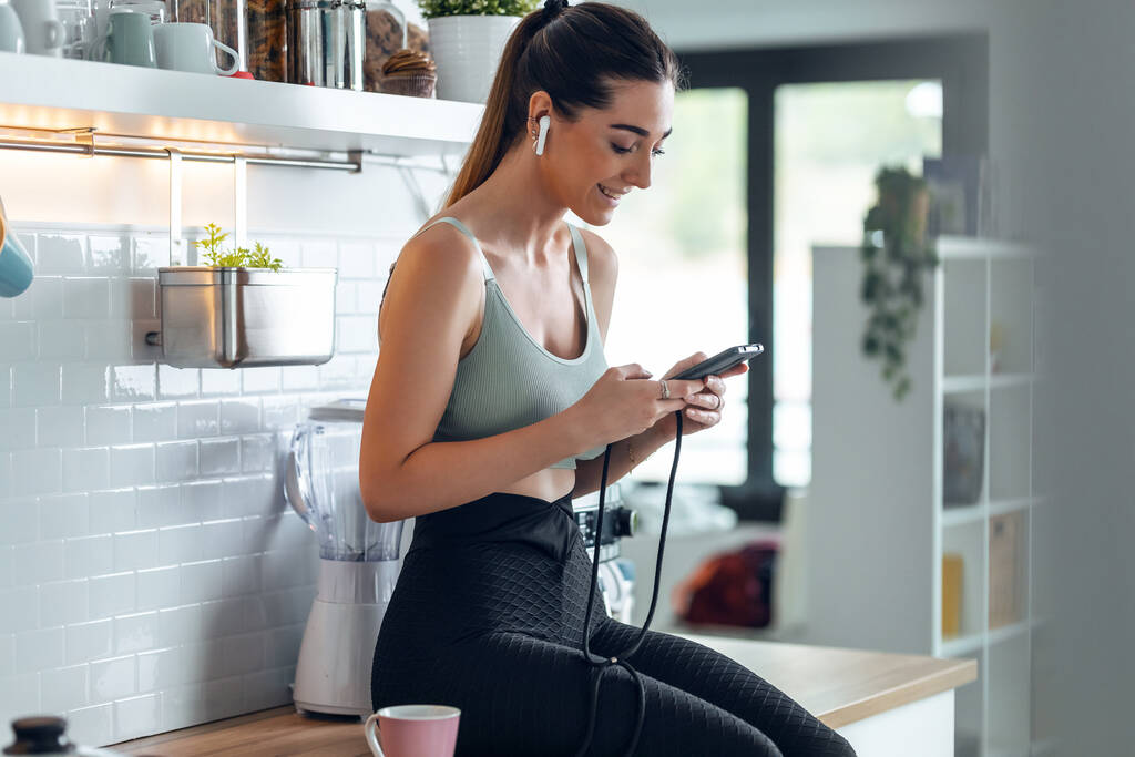 Foto de mujer deportiva escuchando música con su teléfono inteligente, mientras que el café encogimiento en la cocina en casa. - Foto, imagen