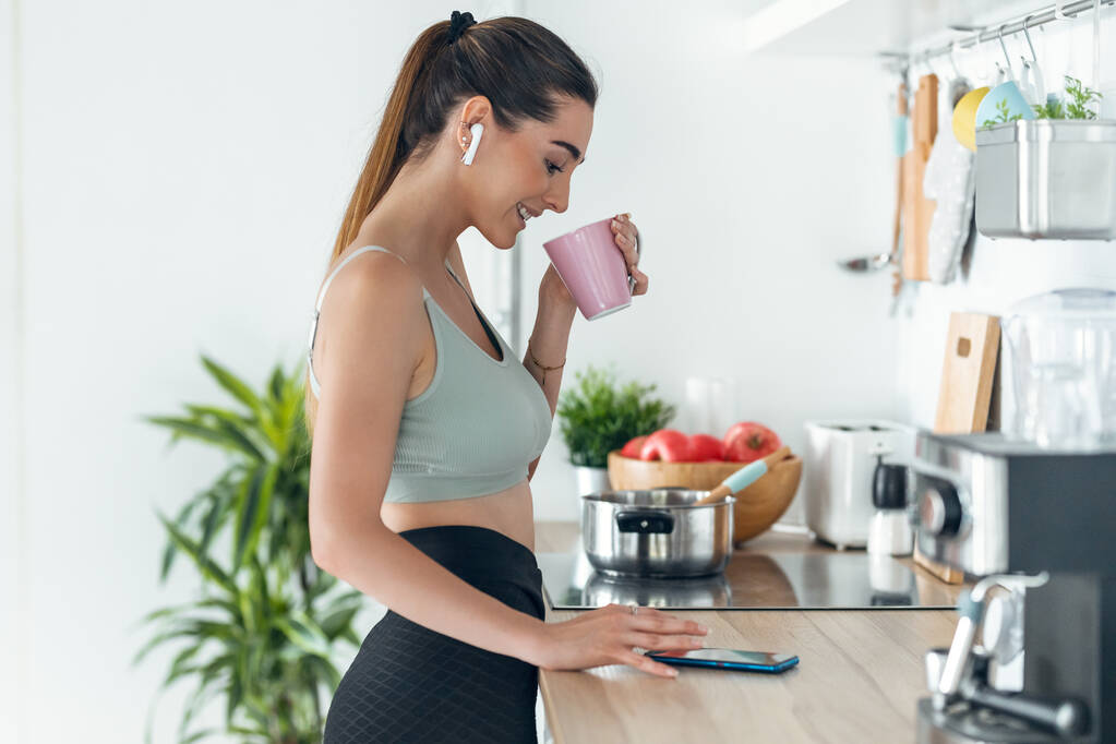 Tiro de mulher esportiva ouvindo música com seu smartphone enquanto bebe café na cozinha em casa. - Foto, Imagem