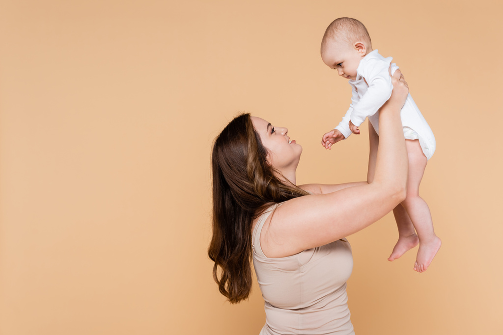 Vista lateral de una mujer sonriente sosteniendo a su hija bebé aislada en beige  - Foto, Imagen