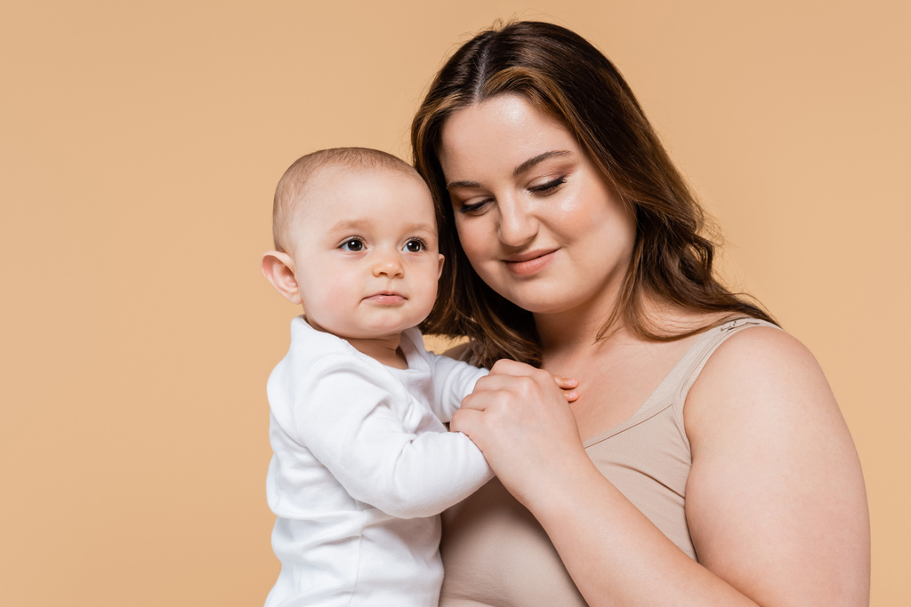 Mujer sonriente con sobrepeso sosteniendo a la hija del bebé aislada en beige  - Foto, imagen