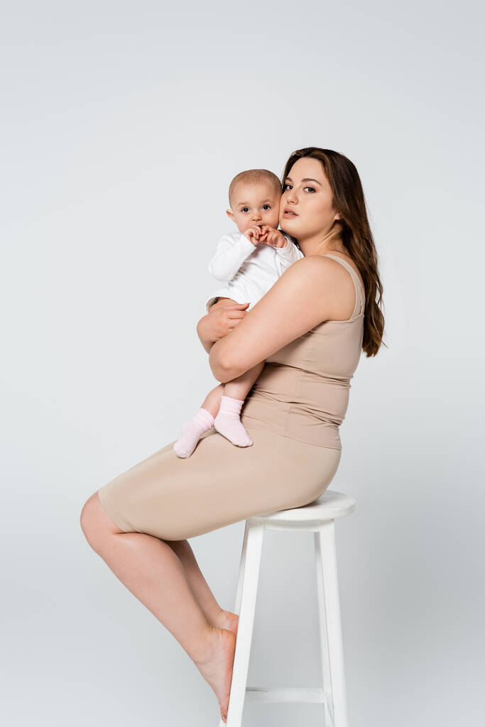 Genç ve cüsseli anne, gri sandalyede izole bir şekilde otururken bebeği kucağına alıyor. - Fotoğraf, Görsel
