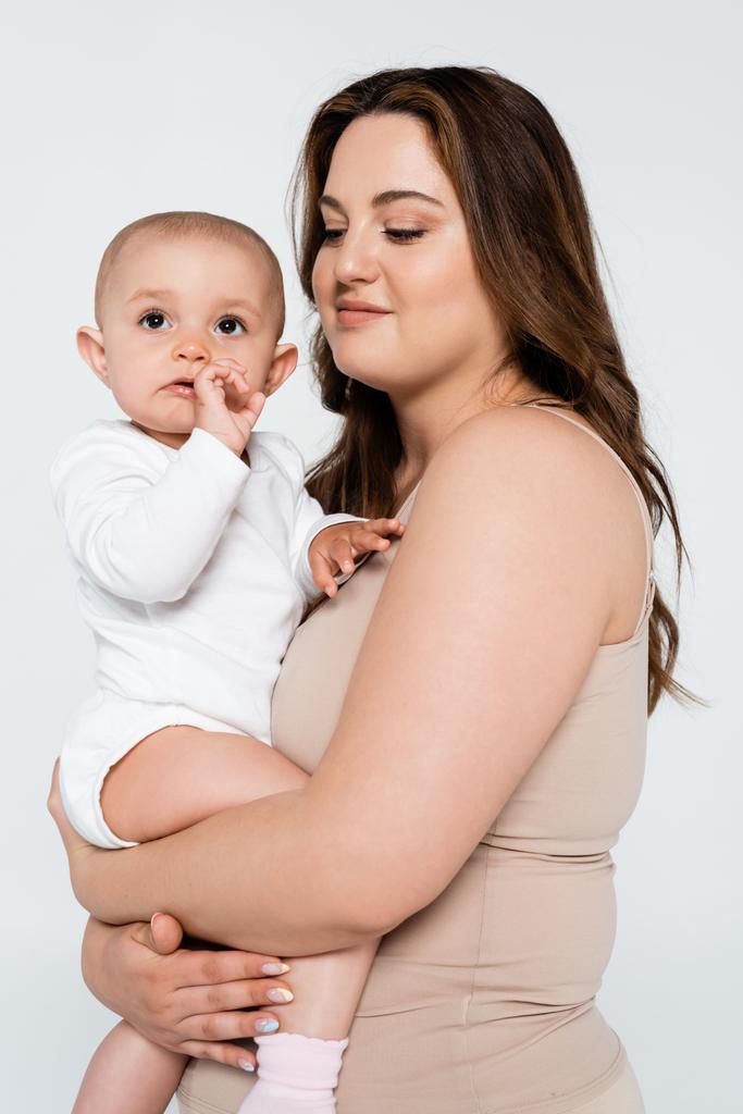 Sonriente madre con sobrepeso abrazo bebé niña aislado en gris - Foto, imagen