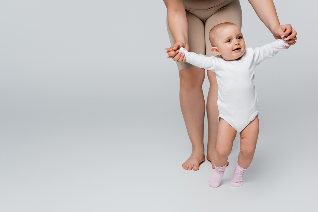 Körper positive Mutter hält Hände von lächelnden Baby-Tochter auf grauem Hintergrund - Foto, Bild