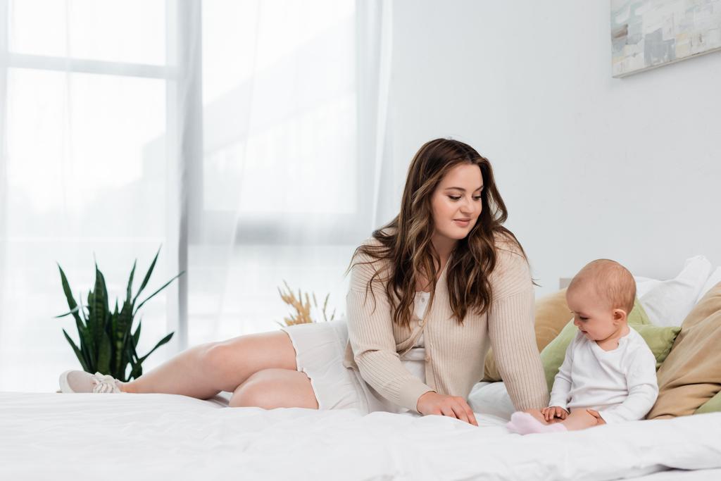 Smiling plus size woman looking at baby daughter on bed  - Photo, Image