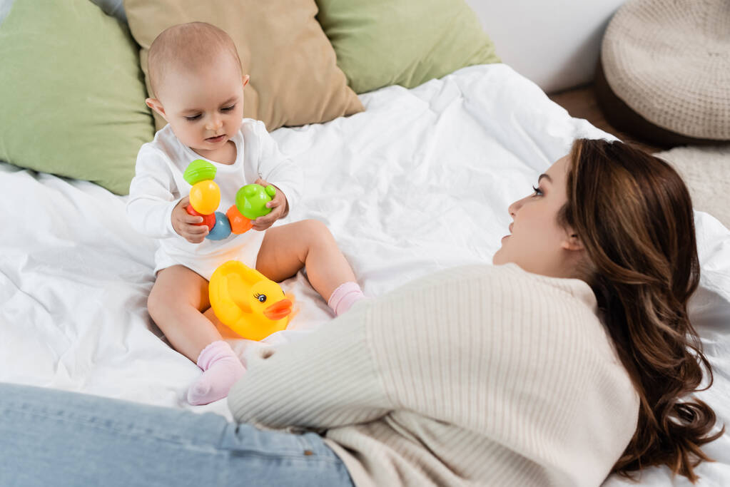 Plus Size Mutter liegt neben Baby Tochter mit Spielzeug auf Bett  - Foto, Bild