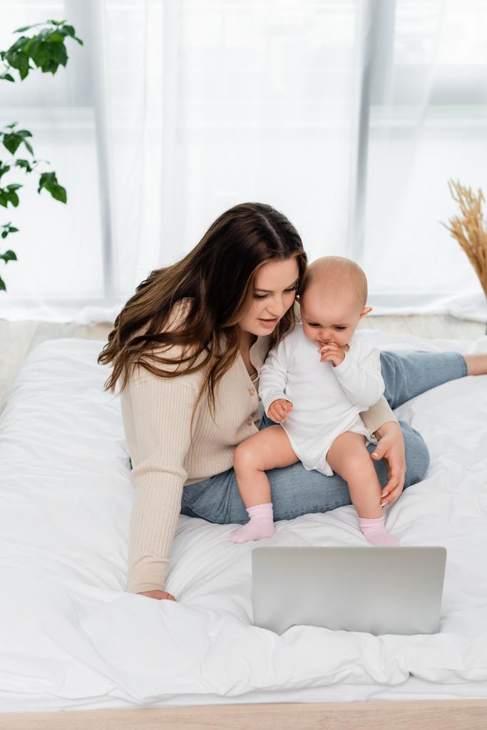 Femme en surpoids tenant bébé fille près d'un ordinateur portable sur le lit  - Photo, image