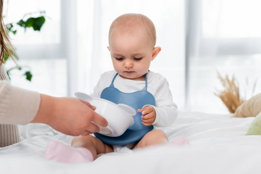 Woman holding plate near baby daughter on bed at home  - Photo, Image