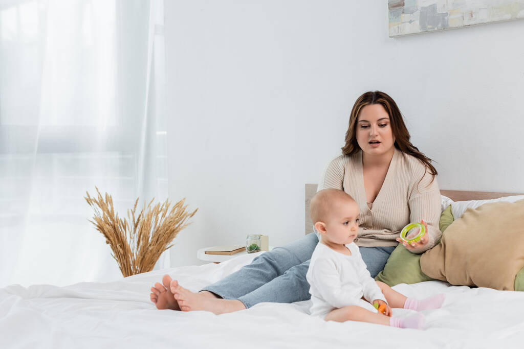 Plus size moeder houden speelgoed in de buurt van baby meisje op bed thuis  - Foto, afbeelding