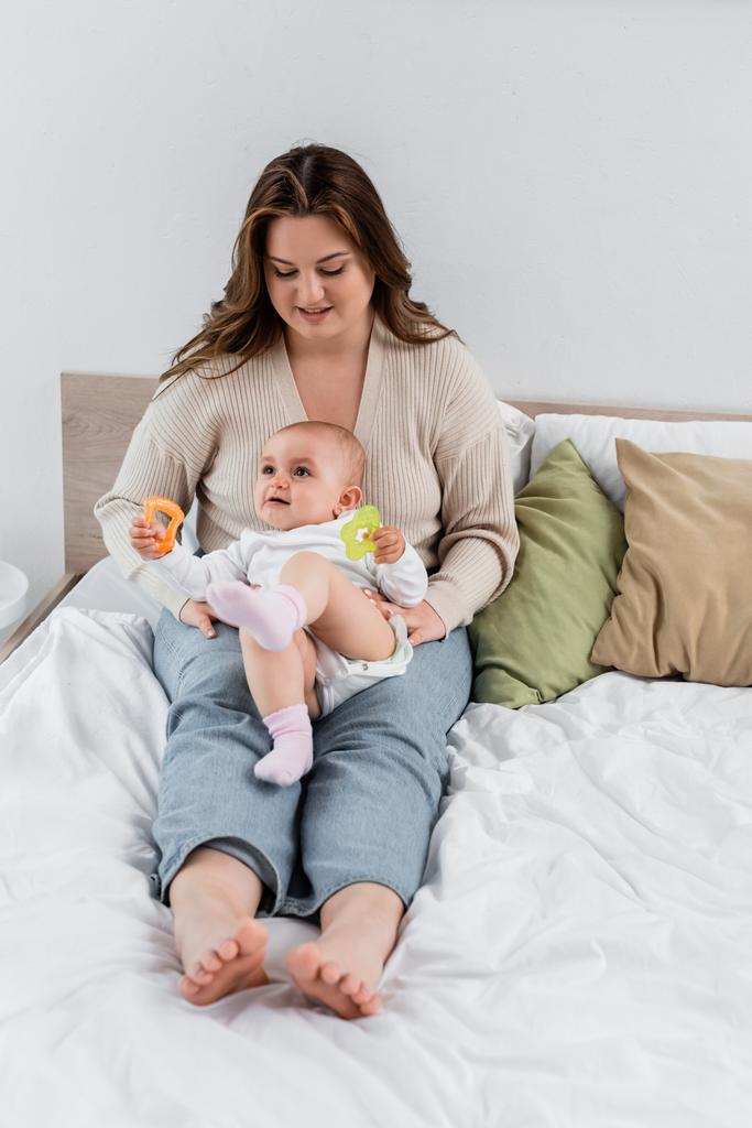 Positive plus size mother looking at baby with toys on bed  - Photo, Image