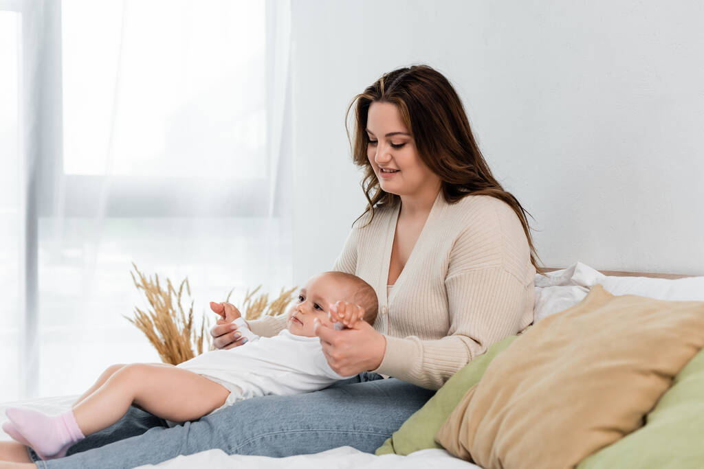 Positivo más tamaño mujer tocando manos de bebé hija en la cama  - Foto, Imagen
