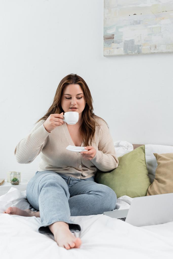 Nuori nainen ylipainoinen tilalla kuppi kahvia lähellä kannettavaa sängyllä  - Valokuva, kuva