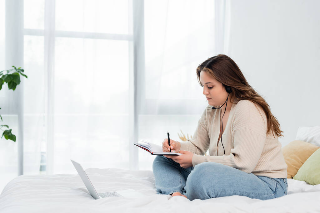 Młody ciało pozytywne freelancer w słuchawki pisanie na notebooku w pobliżu laptopa w sypialni  - Zdjęcie, obraz