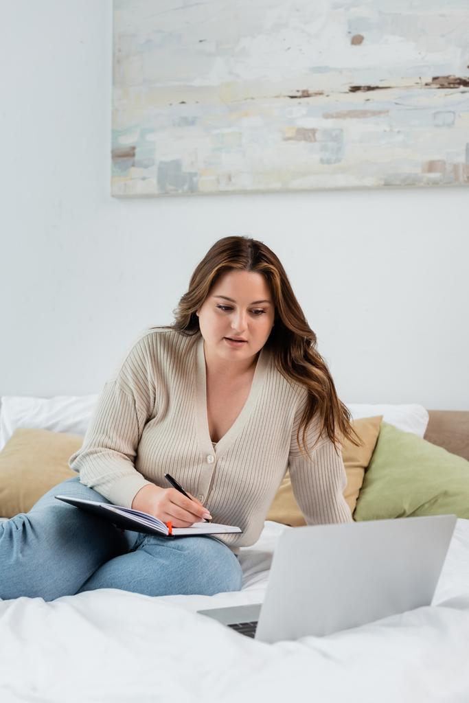 Bruneta žena s nadváhou psaní na notebook v blízkosti rozmazané notebook na posteli  - Fotografie, Obrázek