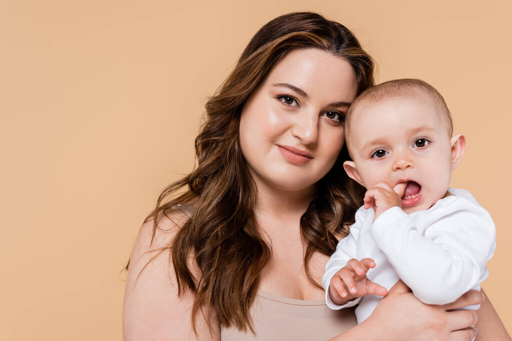 Pretty plus size mother holding baby and looking at camera isolated on beige  - Photo, Image