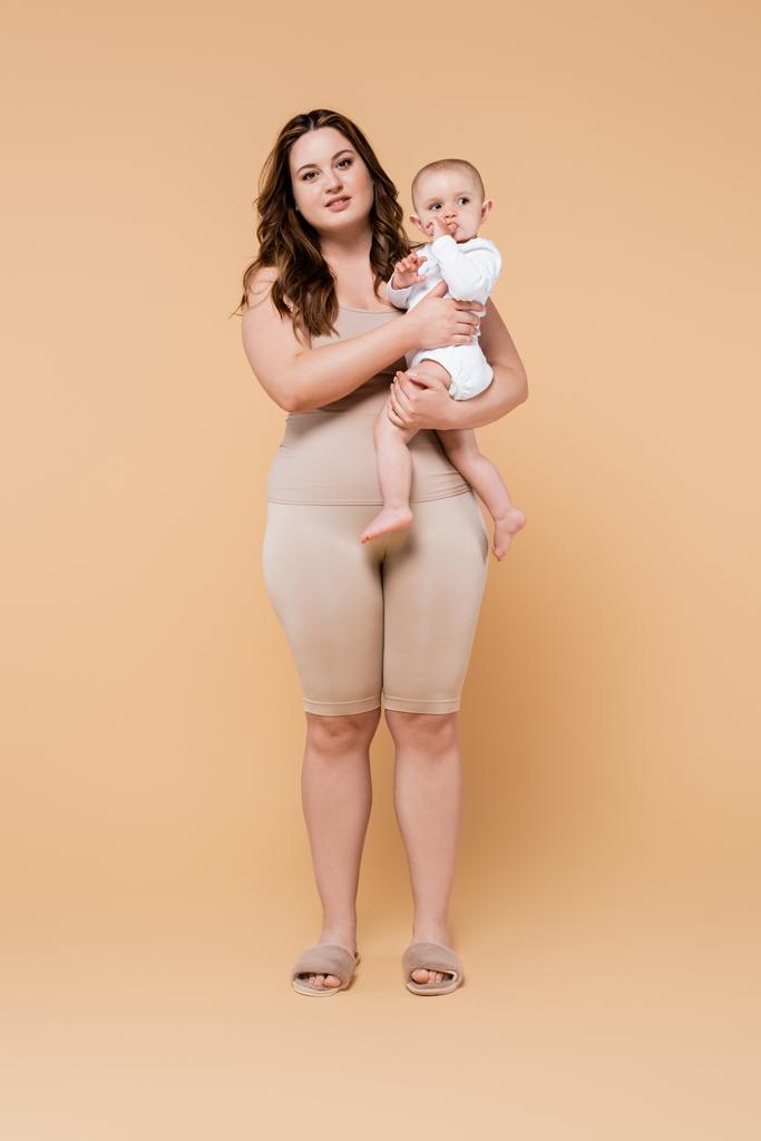 Plná délka těla pozitivní matka drží dceru na béžovém pozadí  - Fotografie, Obrázek