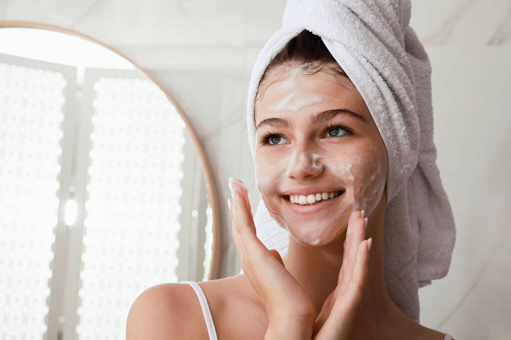 Beautiful teenage girl applying cleansing foam onto face in bathroom, space for text. Skin care cosmetic - Photo, Image