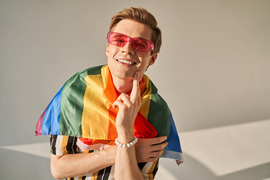 Gay má na tváři vlajku pýchy a růžové brýle, dotýká se tváře a zírá - Fotografie, Obrázek