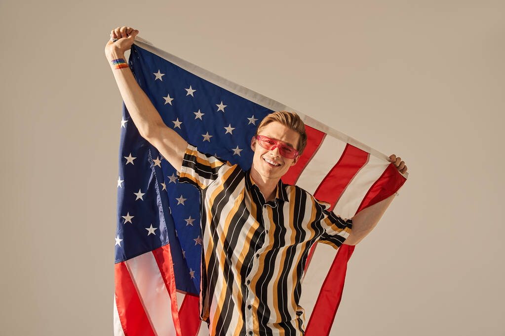 Hombre con el símbolo del arco iris en su brazo sosteniendo la bandera de los estados unidos y sonriendo feliz - Foto, imagen