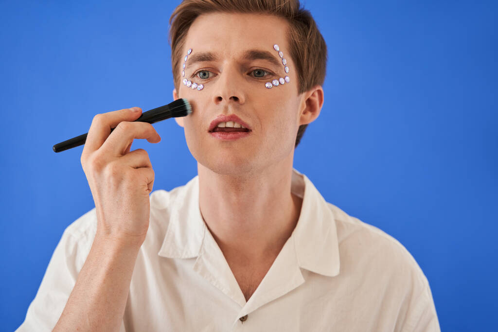 Homem aplicando pó ou blushes e fazendo maquiagem isolada no fundo azul - Foto, Imagem