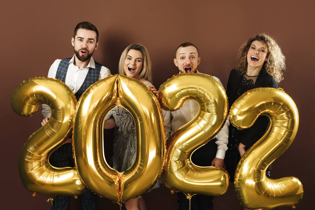 Group of beautiful well dressed party people celebrating arrival of 2022 New Year with a golden balloons on brown background - Photo, Image
