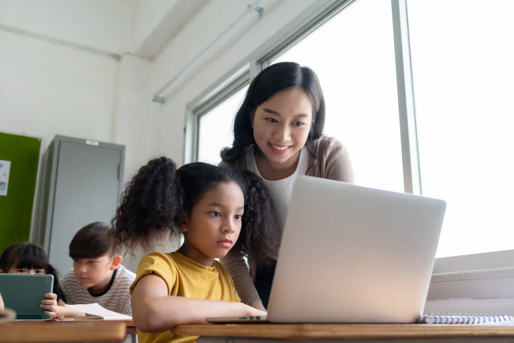 アジアの女性教師は、国際学校の教室でラップトップコンピュータでアフリカ系アメリカ人の女の子を教えています。教育・学習の概念. - 写真・画像