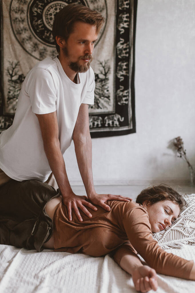 A young Caucasian bearded masseur and a woman doing Thai yoga massage exercises on a mat at home - 写真・画像