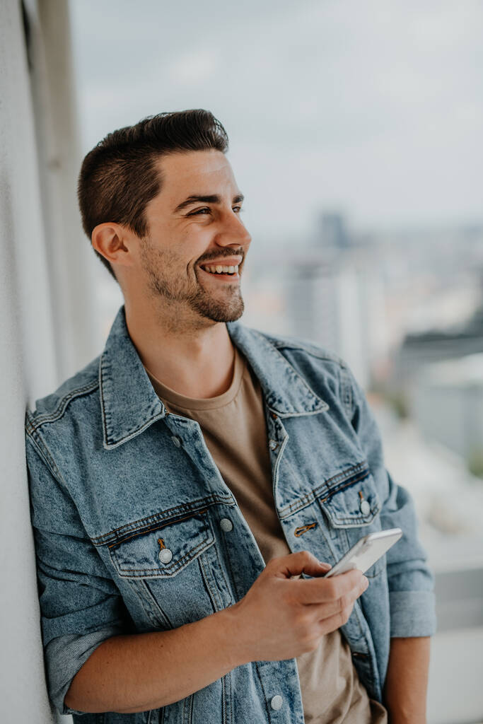 Portrait de jeune homme caucasien heureux avec smartphone debout sur le balcon. - Photo, image