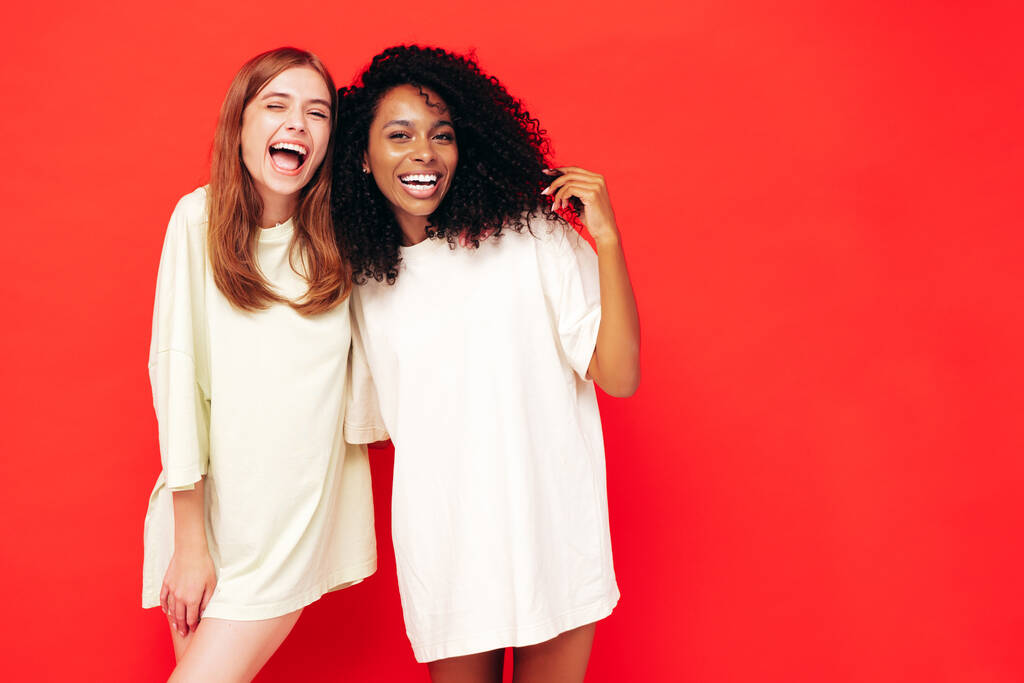 Kaksi nuorta kaunista hymyilevää kansainvälistä hipsterinaista trendikkäissä kesävaatteissa. Seksikkäät huolettomat naiset poseeraavat lähellä punaista seinää studiossa. Positiivisilla malleilla on hauskaa. Ystävyyden käsite - Valokuva, kuva