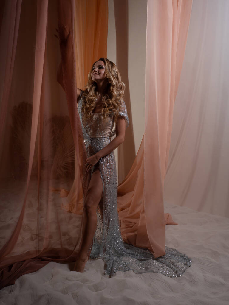 Uma jovem loira elegante em um vestido translúcido brilhante atrás de uma cortina - Foto, Imagem