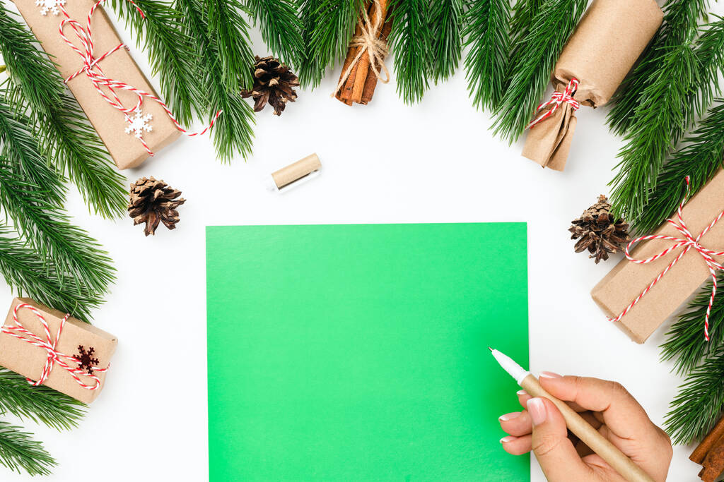 Vánoční přání, zelený dopis s kopírkou a rukou ženy s tužkou na bílém stole a ozdobou z borovicových větví s dárky - Fotografie, Obrázek