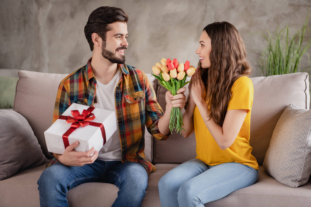Foto von schönen attraktiven jungen Liebhabern gekleidet casual Kleidung geben Geschenk Box Tulpen lächeln drinnen Loft-Wohnung - Foto, Bild