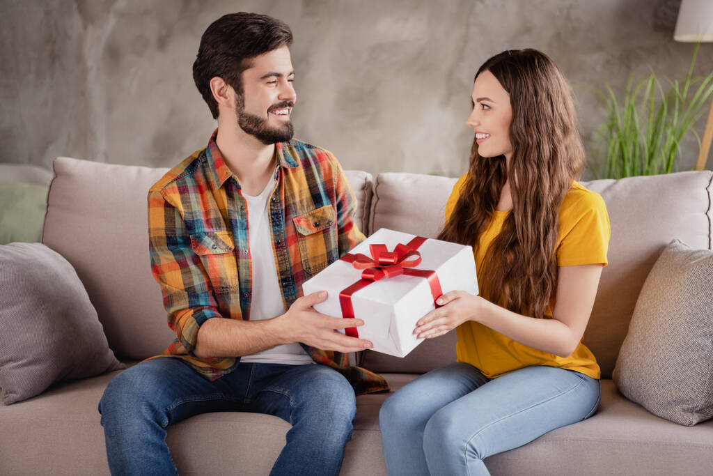 Foto de hermosa pareja joven dulce usar trajes casuales sonriendo presentando caja de regalo habitación interior de la casa - Foto, imagen