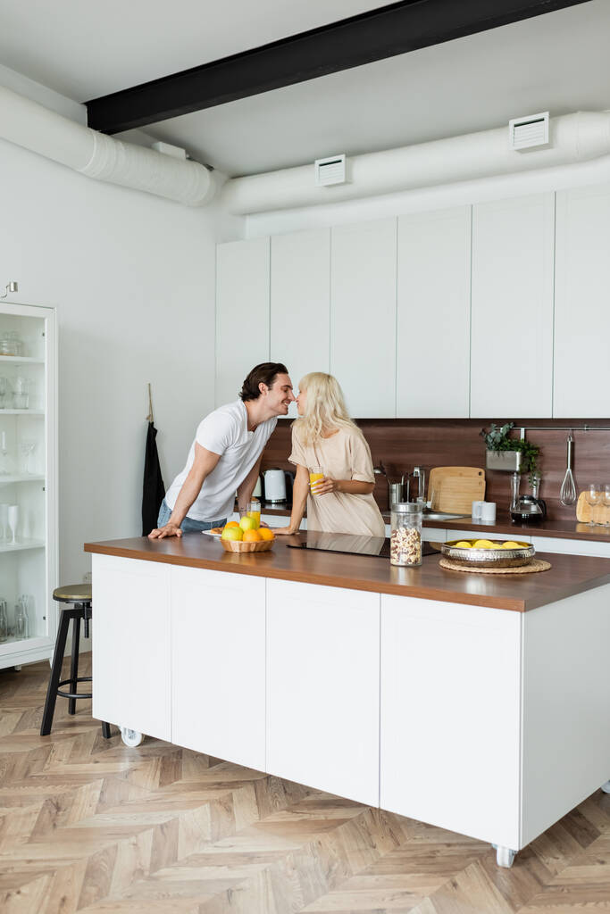 homem feliz beijando namorada alegre com vidro de suco de laranja na cozinha - Foto, Imagem