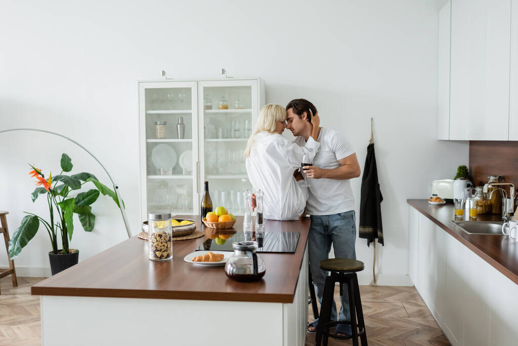 νεαρό ζευγάρι με ποτήρια κρασί αγκαλιάζει στη σύγχρονη κουζίνα  - Φωτογραφία, εικόνα