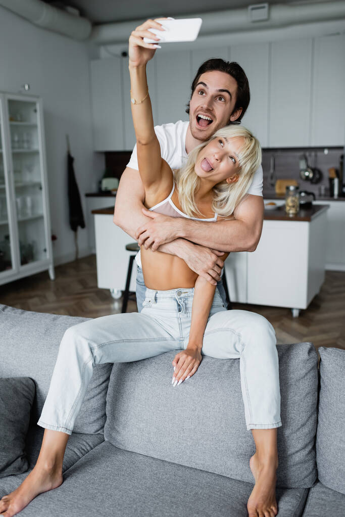 rubia mujer sobresaliendo lengua mientras toma selfie con feliz novio en sala de estar  - Foto, imagen