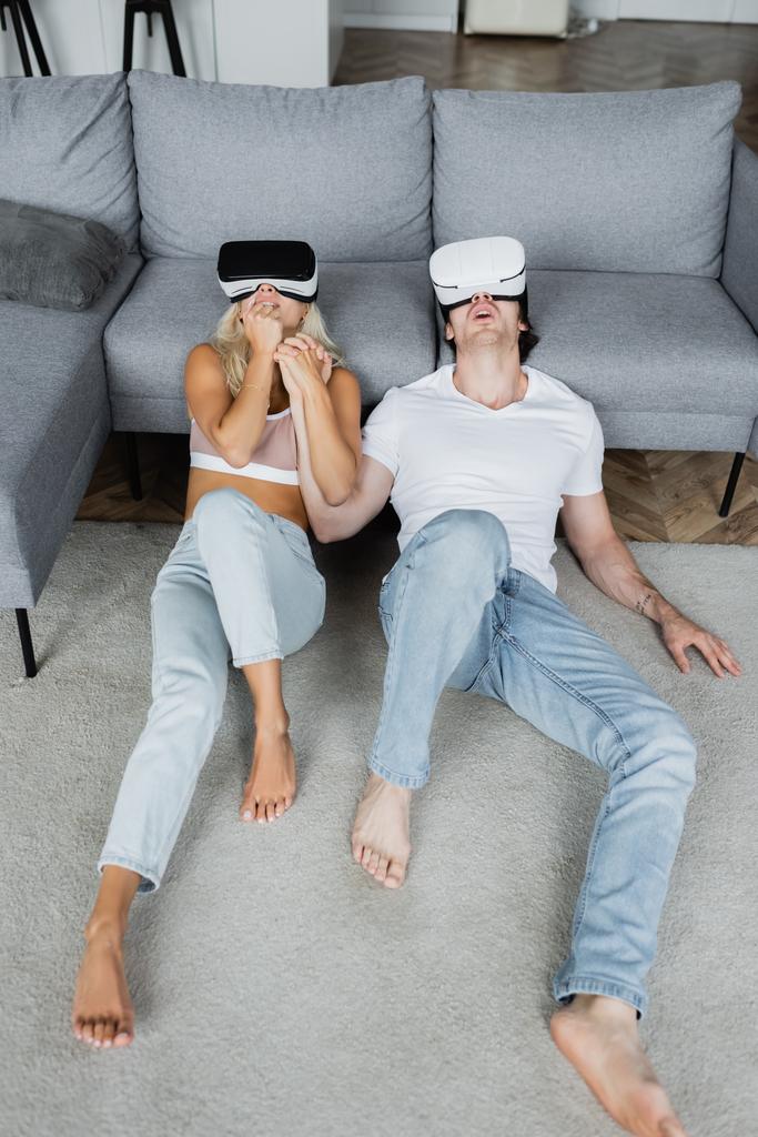 σοκαρισμένο ζευγάρι σε ακουστικά VR κάθεται στο χαλί κοντά γκρι καναπέ - Φωτογραφία, εικόνα