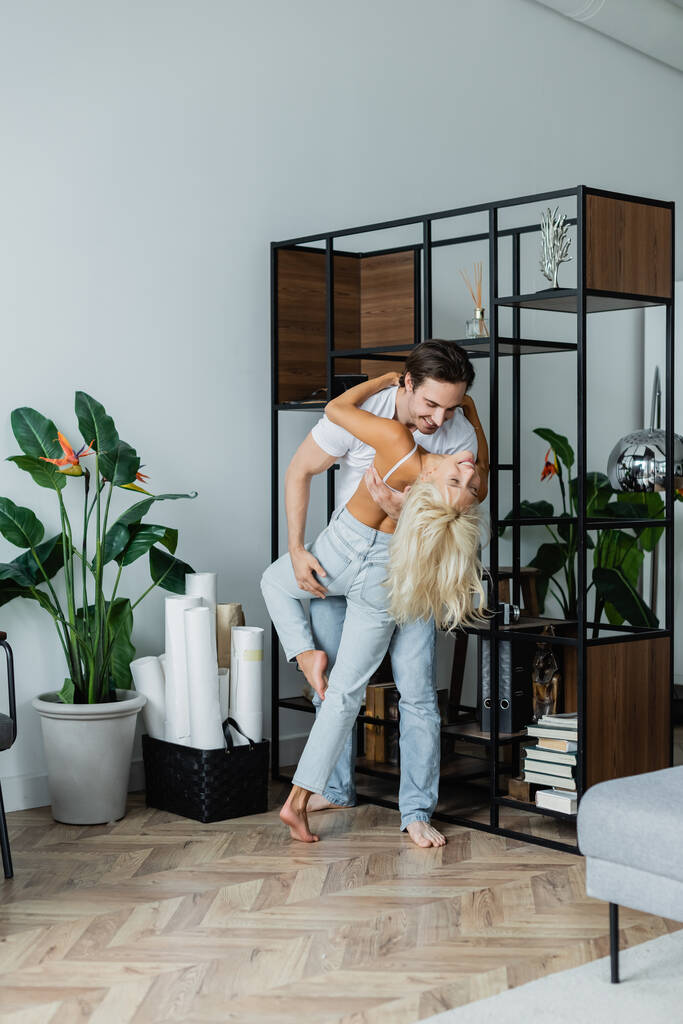 full length of happy man bending cheerful woman while dancing near rack in living room - Photo, Image