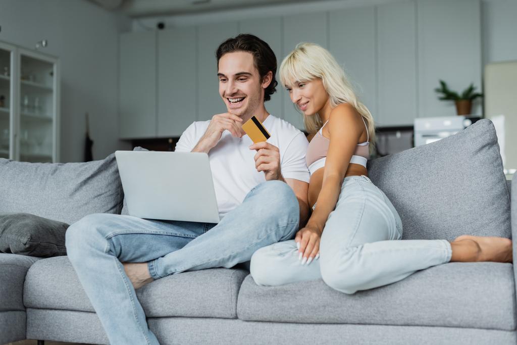 fröhlicher Mann mit Kreditkarte in der Nähe seiner Freundin, während er auf Laptop schaut und online einkauft  - Foto, Bild